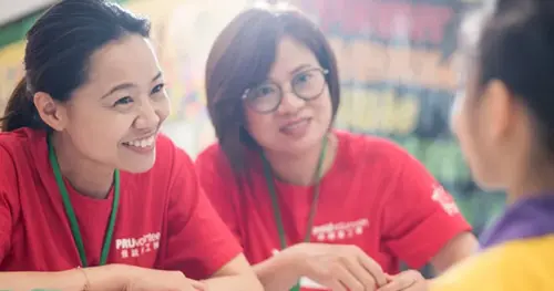 Two women in red shirts with name tags smile and talk to a third person, who is slightly blurred in the foreground. The setting appears to be informal and friendly.