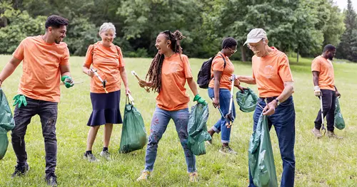 Volunteers in orange shirts clean a park, smiling as they carry green trash bags and use litter pickers.
