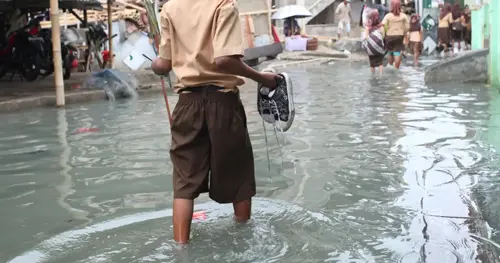A child in a brown school uniform holds shoes and walks through knee-deep floodwater on a street, while other people wade in the background amid submerged buildings and debris.