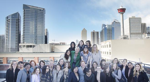 A large group of women pose and smile together on a rooftop, celebrating International Women’s Day with the Calgary Tower and modern city buildings visible in the background under a partly cloudy sky.