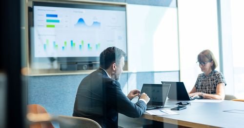 2 employees working on finance with an analytics dashbord in a tv