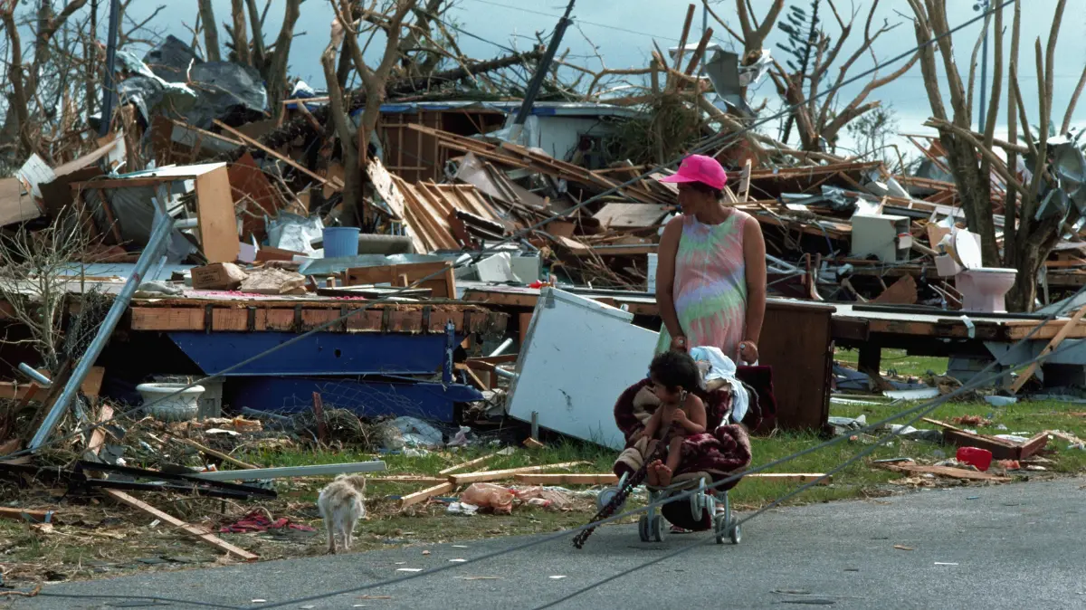 A woman pushes a child in a stroller past destroyed homes and debris, with broken trees and scattered belongings visible.