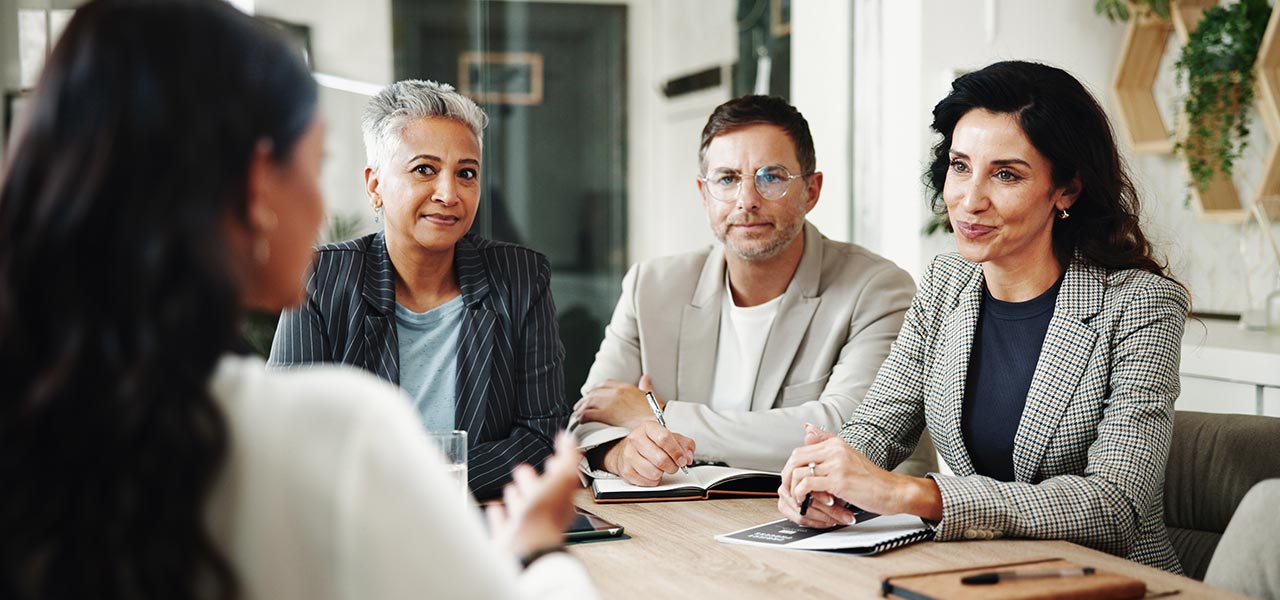 Three professionals sit at a table, attentively listening and taking notes as they interview a woman whose back is to the camera. The setting is a modern office with bright natural light.