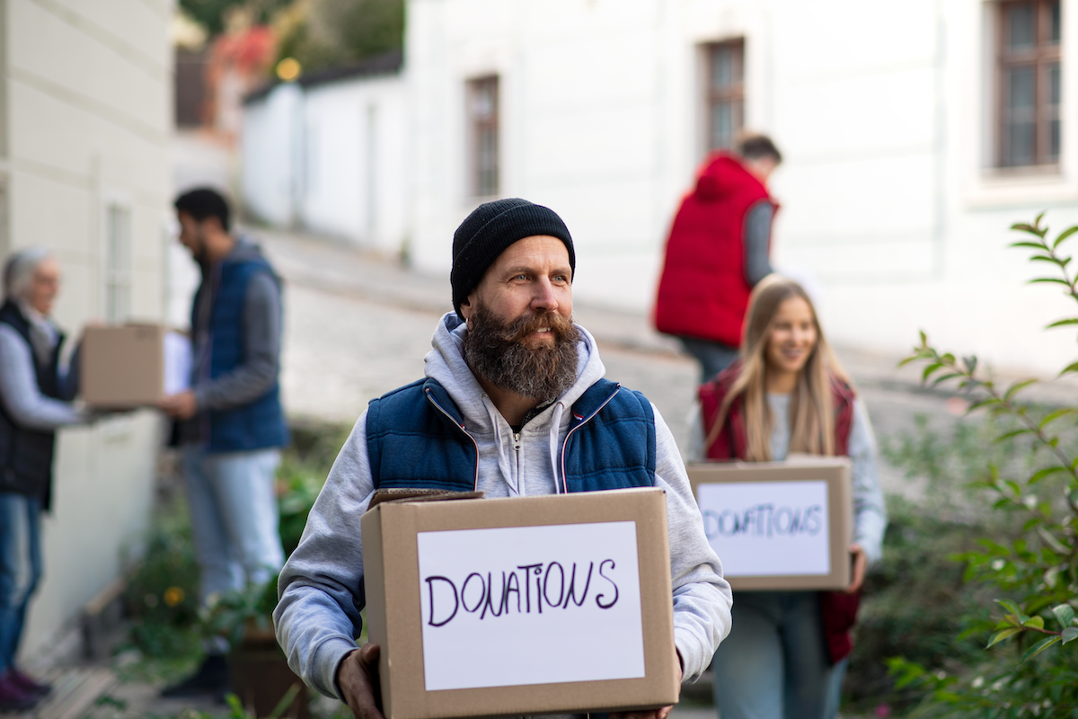 A bearded man in a beanie carries a box labeled Donations. Behind him, several people also carry donation boxes outdoors, forming a line and passing boxes along beside a building.