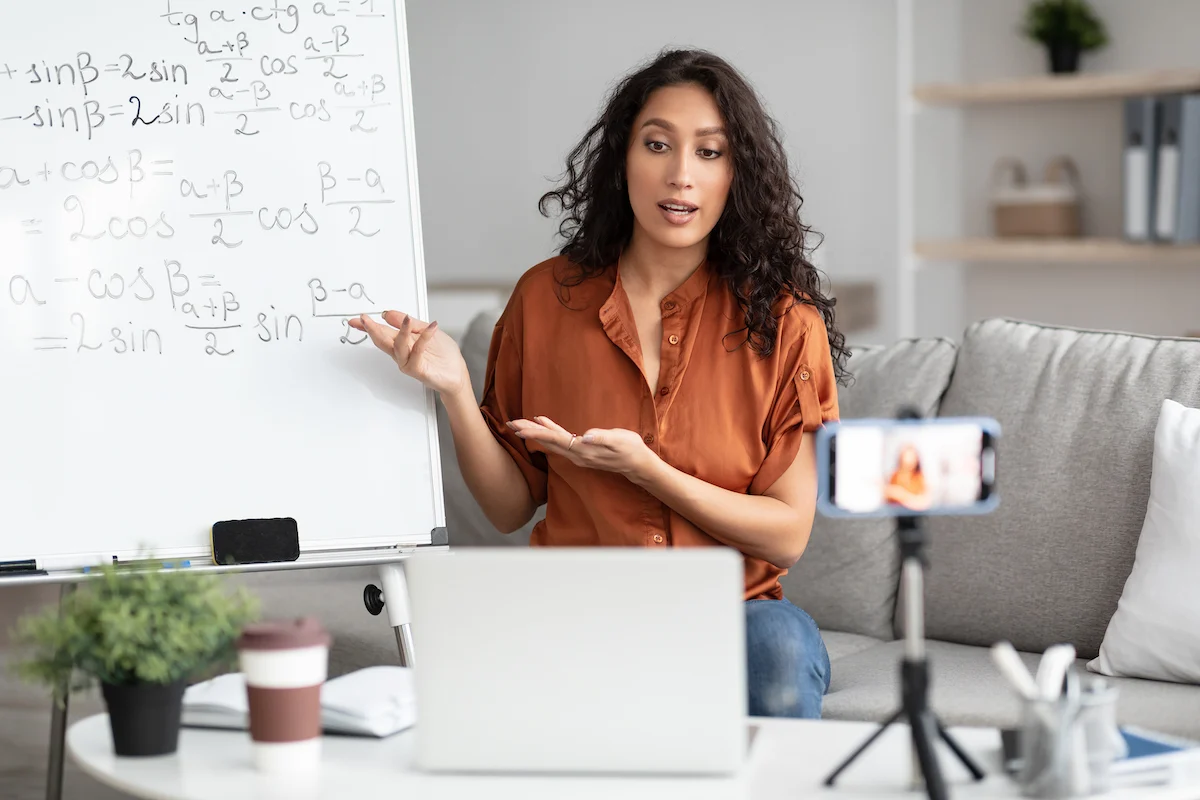 A woman sits on a couch in front of a laptop, pointing at math equations on a whiteboard while speaking to a smartphone camera set up on a tripod, suggesting she is teaching or tutoring online.