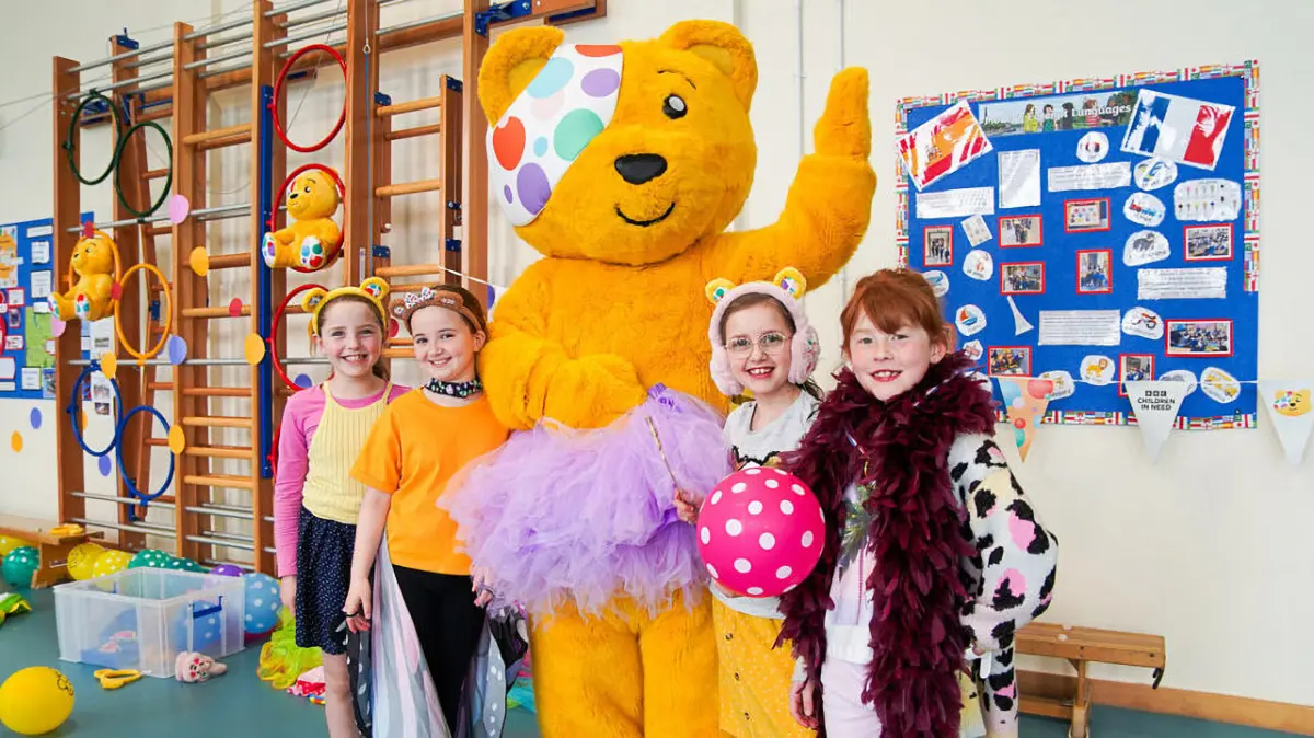 Four children dressed in colorful costumes stand beside a person in a large Pudsey Bear costume, smiling in a decorated room with balloons, posters, and children’s artwork on the wall.