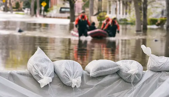 Four sandbags line a barrier in the foreground of a flooded street, while rescue workers in orange jackets transport people in a small inflatable boat in the background.