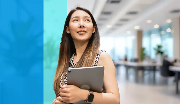 A woman with long hair, wearing a sleeveless patterned top and a smartwatch, smiles while holding a tablet in a modern, bright office space with desks and plants in the background.