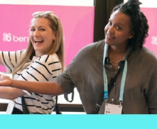 Two women smiling and posing together in front of a pink background with ben part of a logo visible. One woman wears a striped shirt and the other a gray shirt with a lanyard.