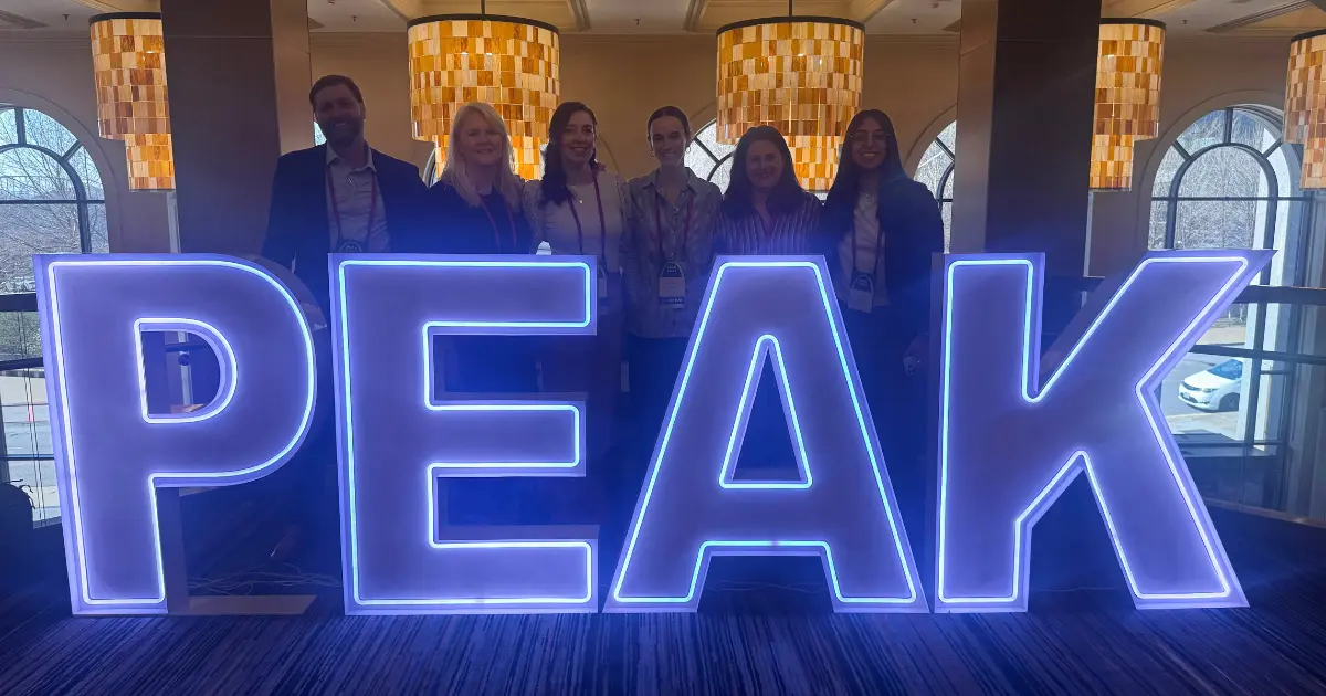 Seven people stand behind large, illuminated letters spelling PEAK in a brightly lit indoor space with hanging lamps and arched windows in the background.