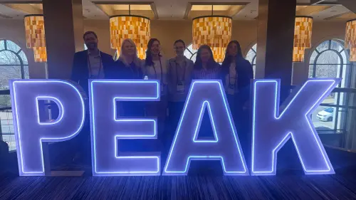 Seven people stand behind large, illuminated letters spelling PEAK in a brightly lit indoor space with hanging lamps and arched windows in the background.