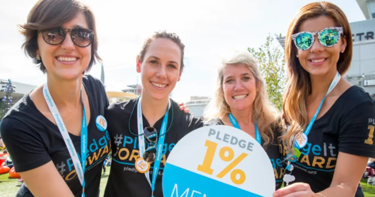 Four smiling women wearing black “#PledgeItForward” shirts and blue lanyards hold a sign reading Pledge 1%. They are outdoors on a sunny day, posing together for a group photo.
