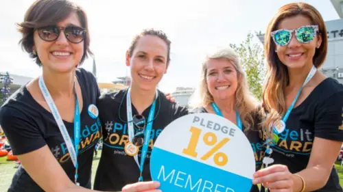 Four smiling women wearing black “#PledgeItForward” shirts and blue lanyards hold a sign reading Pledge 1%. They are outdoors on a sunny day, posing together for a group photo.