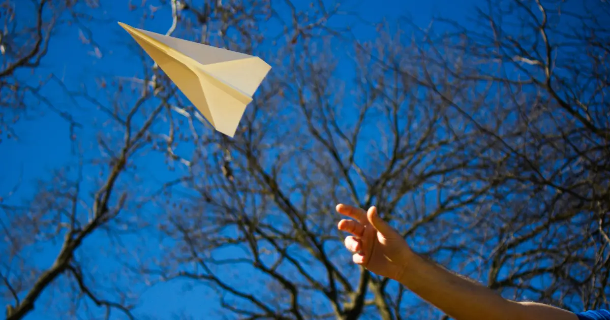 A hand throws a paper airplane into the air on a clear day, with leafless tree branches and a bright blue sky in the background.