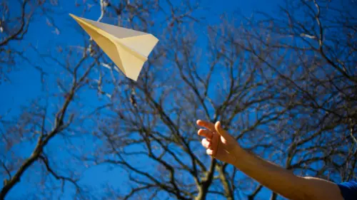 A hand throws a paper airplane into the air on a clear day, with leafless tree branches and a bright blue sky in the background.