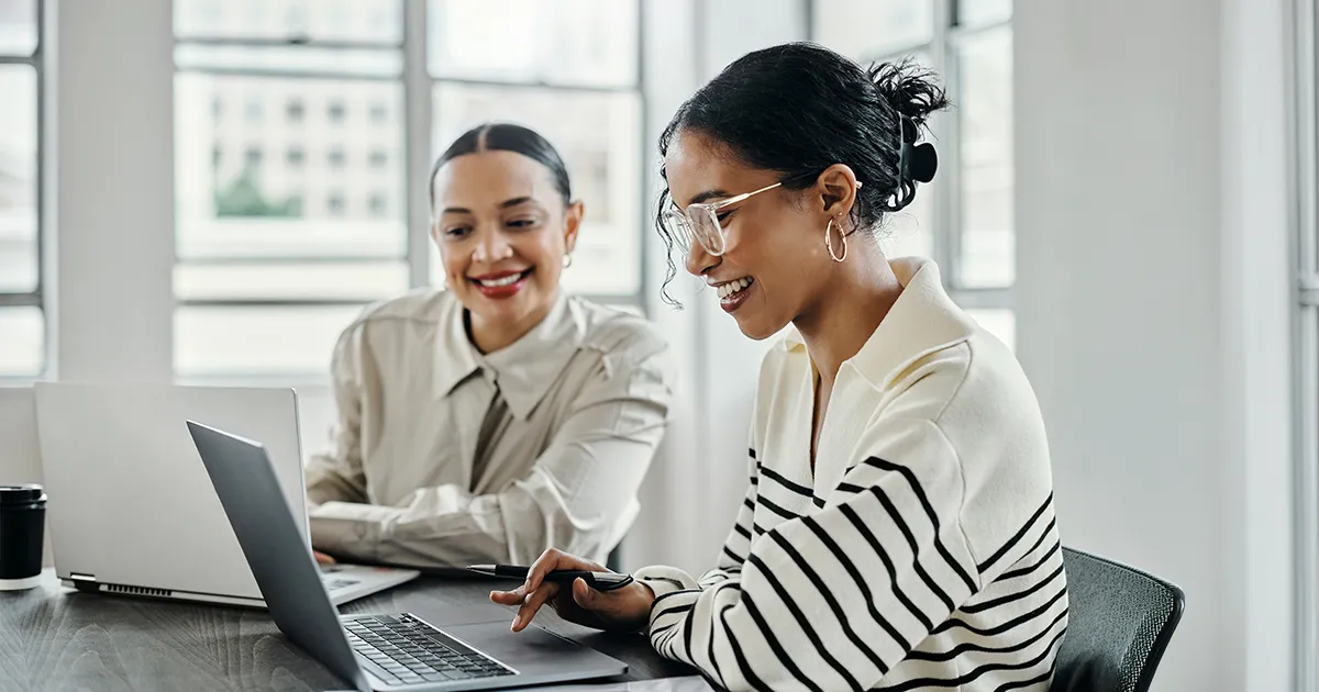Two coworkers at a bright office on their laptops reviewing talking and reviewing their work.