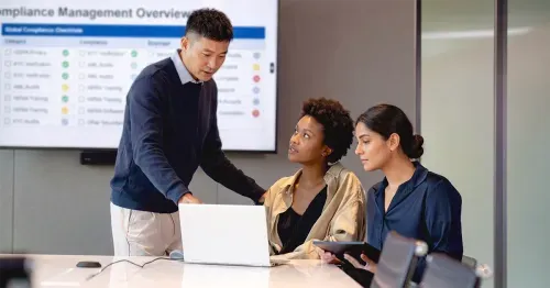 Corporate colleagues discussing compliance in a boardroom with a compliance checklist on a tv screen behind them.