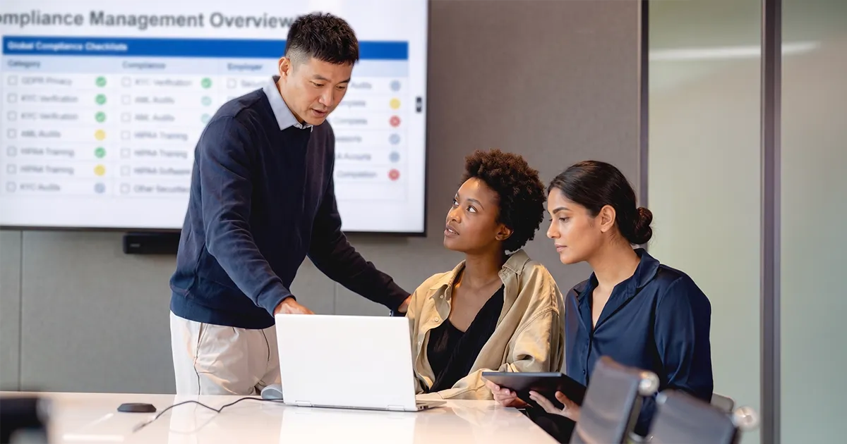 Corporate colleagues discussing compliance in a boardroom with a compliance checklist on a tv screen behind them.