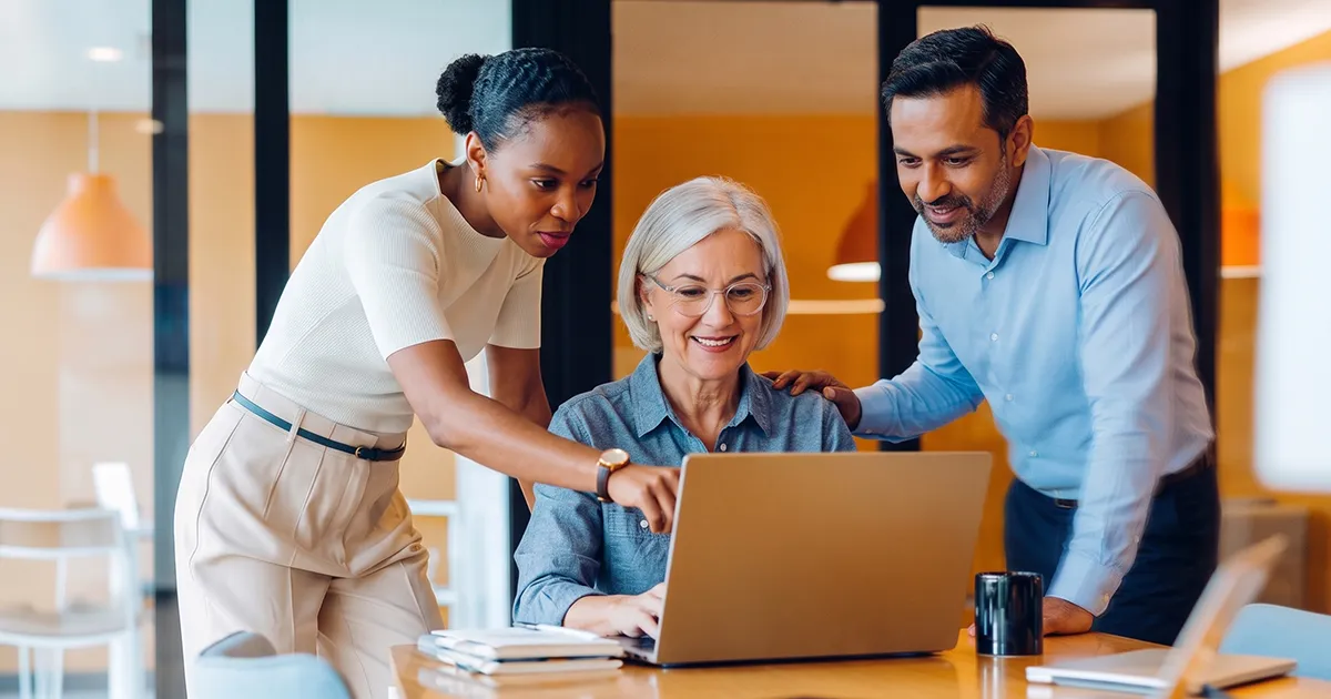 3 coworkers surrounding and looking at a laptop in a bright modern office.