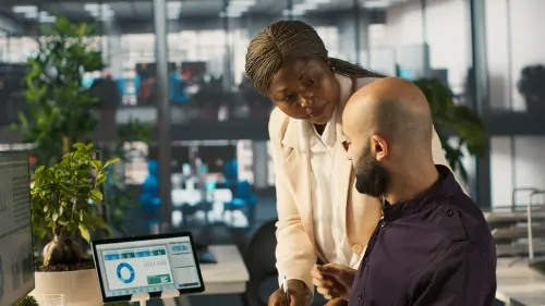 A woman in a light blazer leans over a colleague in a dark shirt at his desk, both looking at a document as he writes with a pen, with a monitor showing colorful analytics dashboards beside them in a modern office