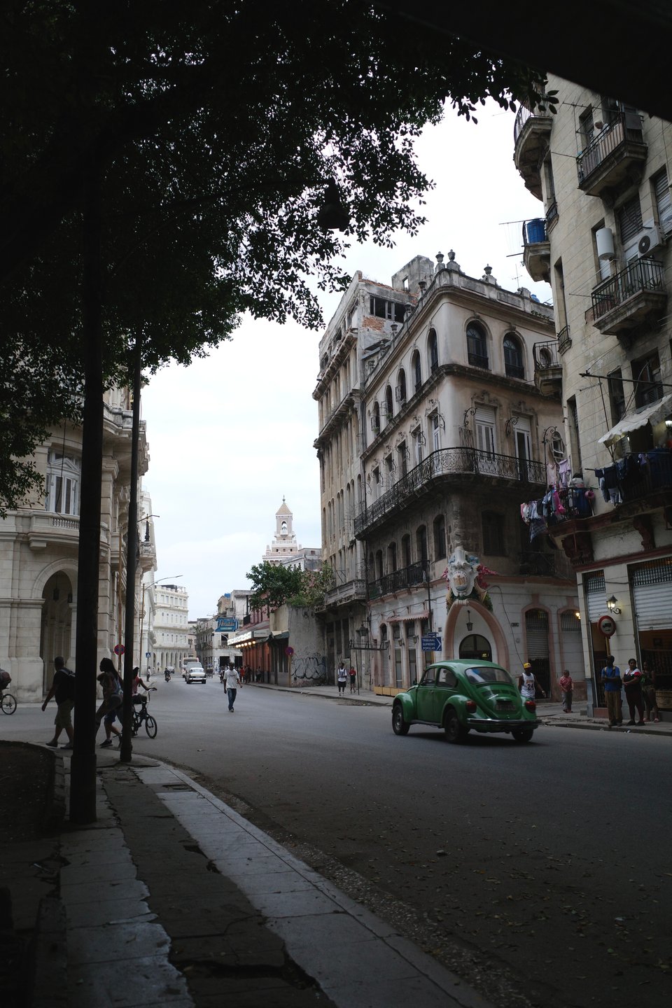 Street view of Havana, featuring classic colonial buildings, a green vintage car driving past, and people walking and cycling along the road.