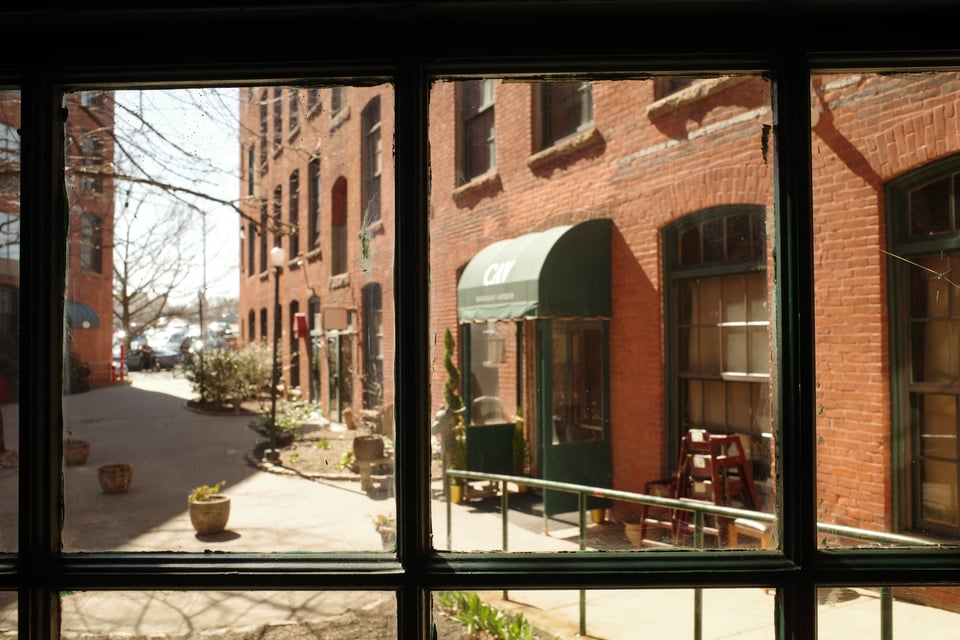 View through a multi-pane window into a quiet brick alleyway with green awnings, potted plants, and warm sunlight filtering in.