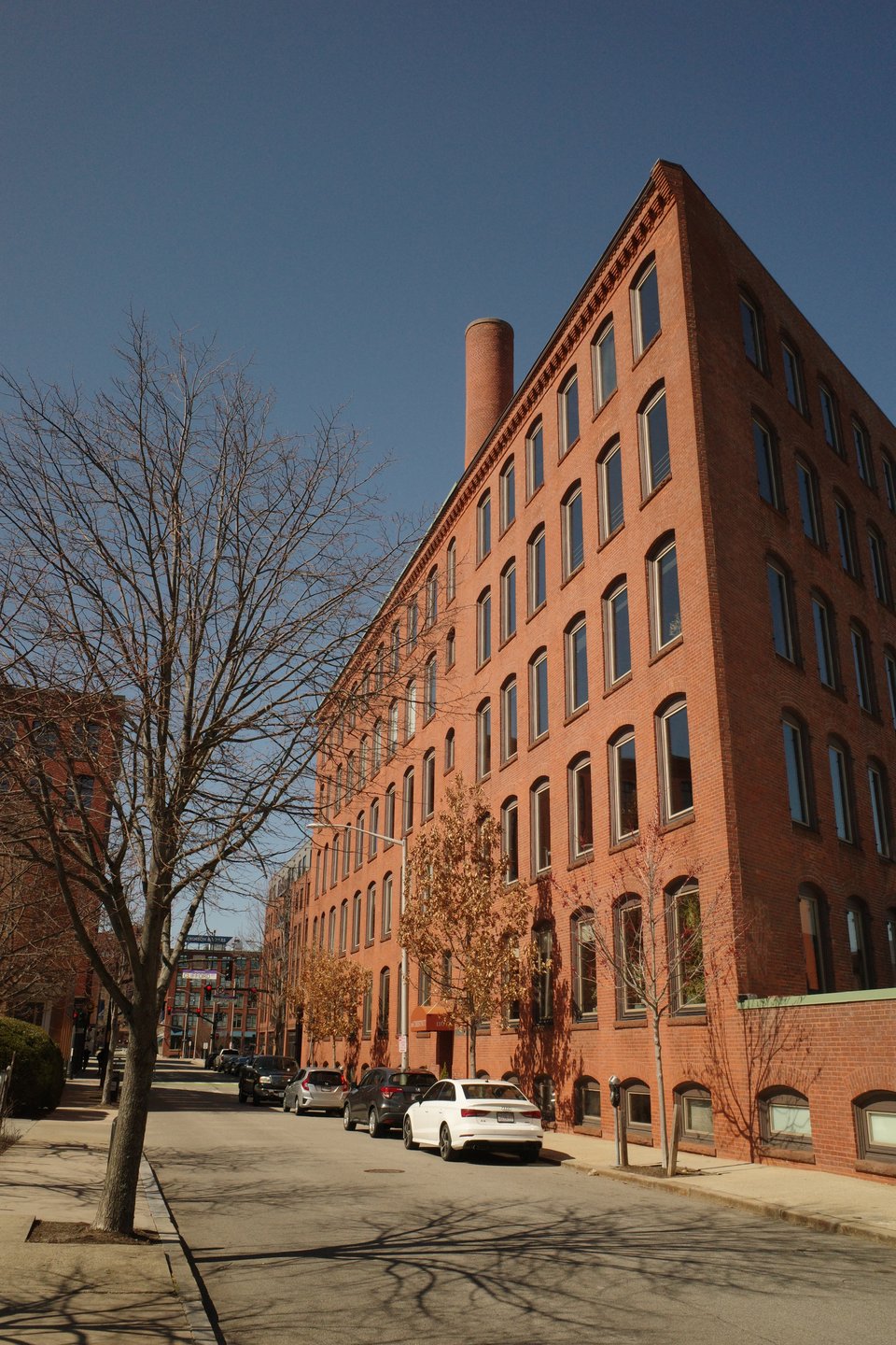 A corner view of a tall brick building under a deep blue sky, with bare trees and cars lining the street.