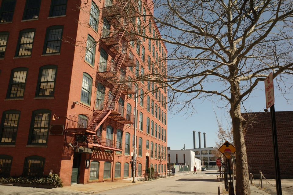 A brick building with fire escapes and arched windows, with factory smokestacks in the distance.