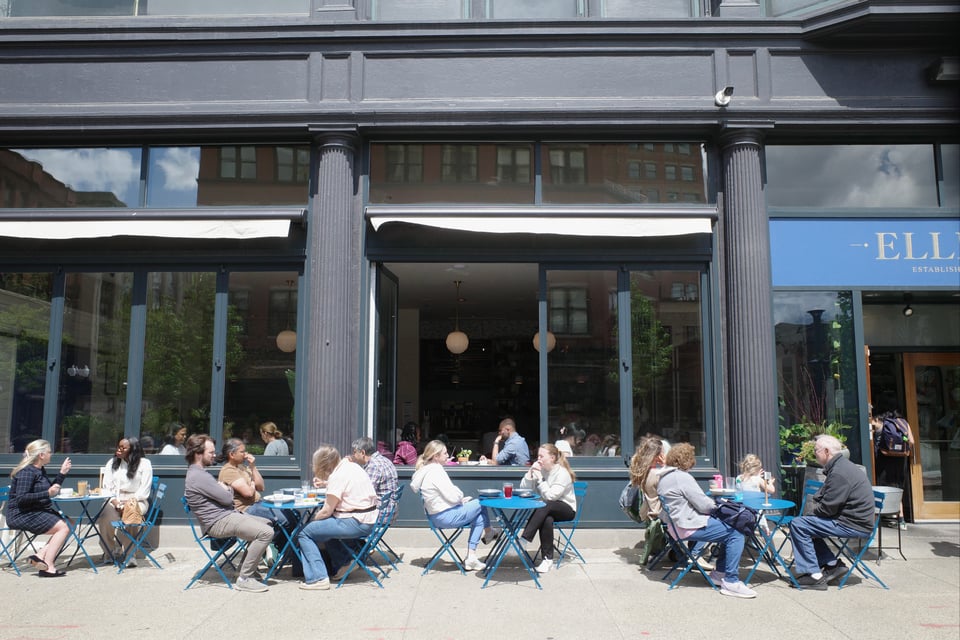 People sit at blue tables outside a café, enjoying sunny but slightly windy weather.
