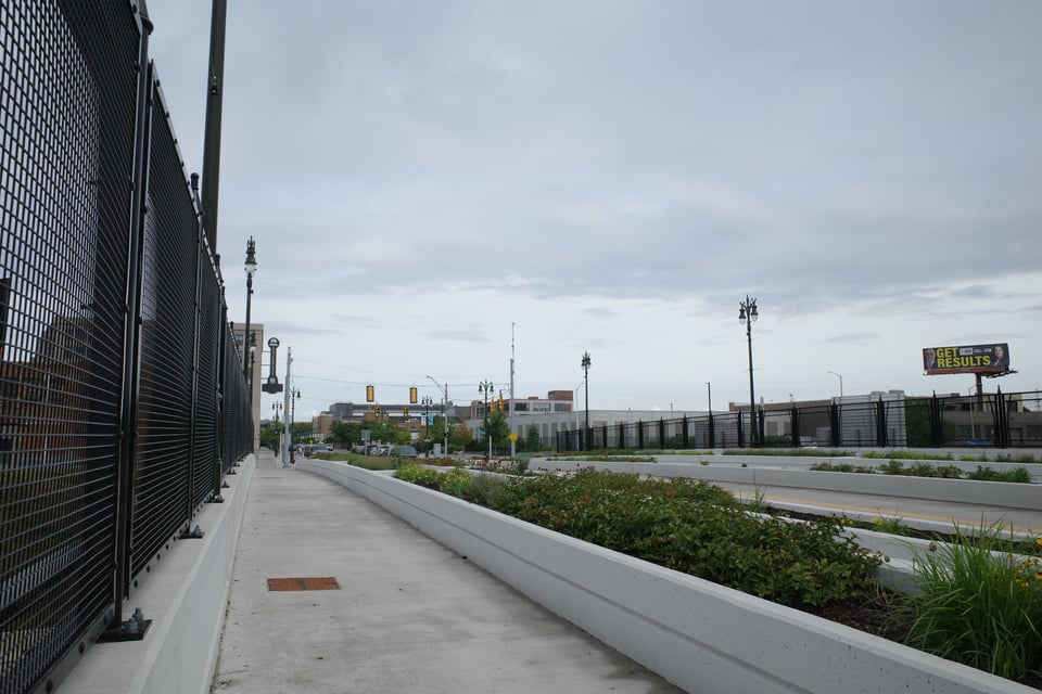 Wide sidewalk and planted median along a highway overpass on a cloudy day, with a black fresh-looking metal fence on the left.