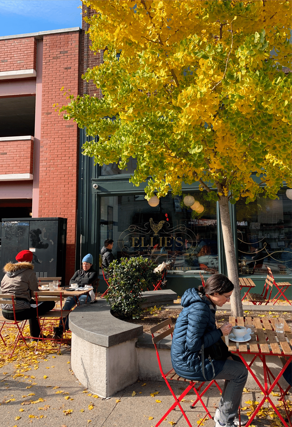Outdoor tables in front of the old Ellie's, with people bundled in coats and hats sitting under a bright yellow tree, the café sign “Ellie’s” visible on the window.