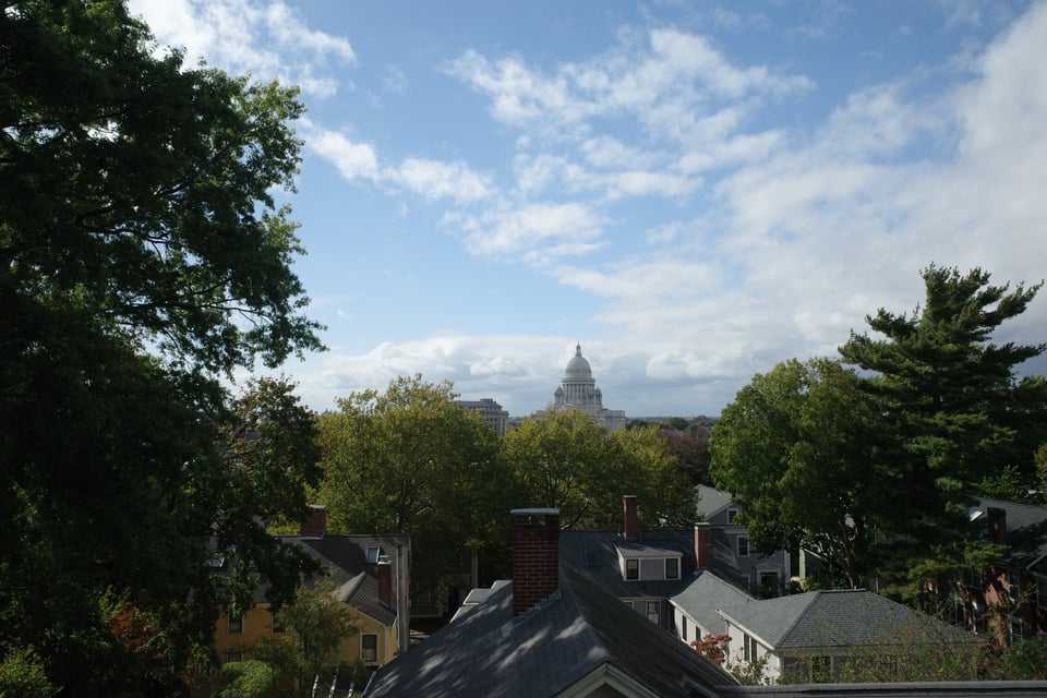 Rooftops of historic Providence houses framed by trees, with the white Rhode Island State House dome rising in the distance under a partly cloudy sky.