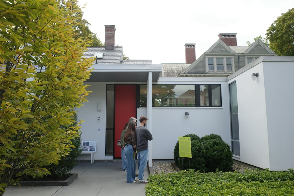 Minimal white modern home with a tall red front door and broad horizontal windows; several tour attendees stand and talk in front of the entrance.