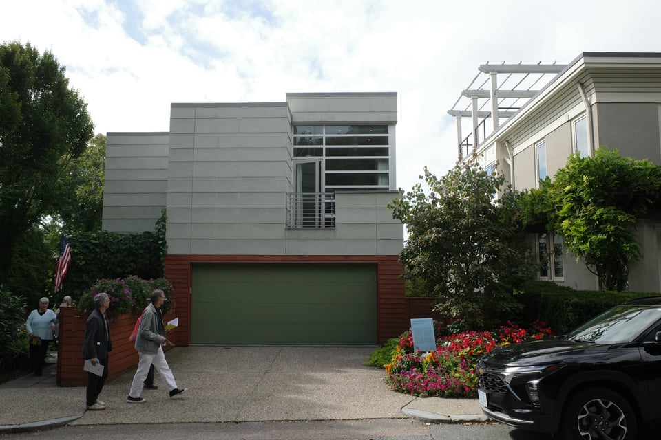 Two-story modern home with light-gray paneling and a green garage door, landscaped with flowers; people from a house tour walk along the driveway.