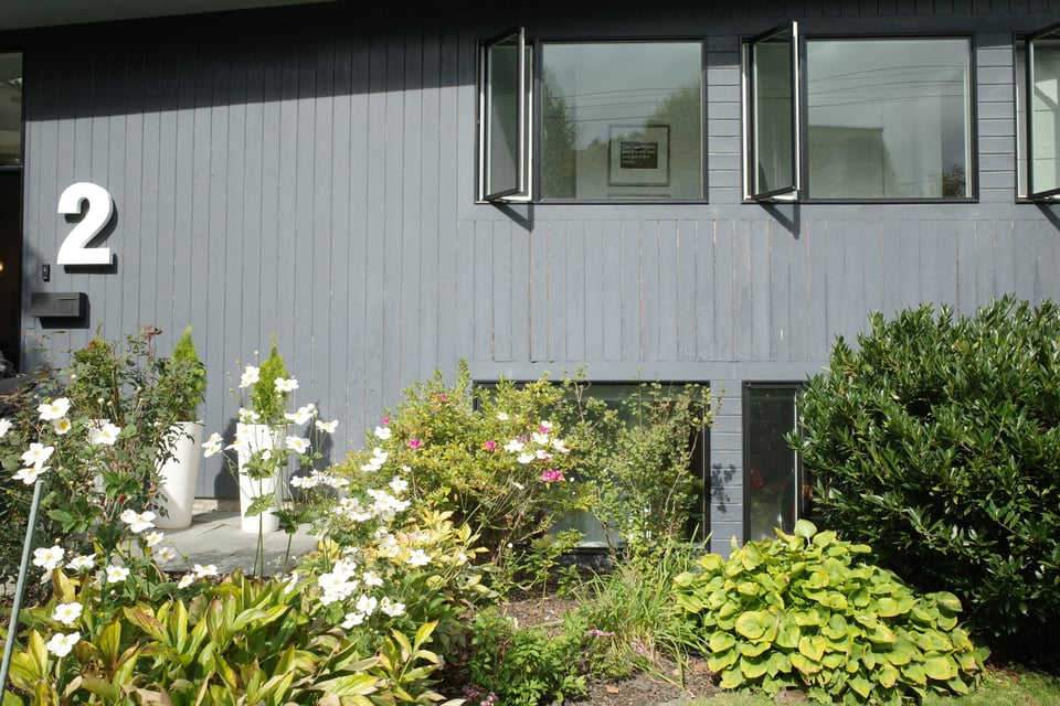 Front of a gray, mid-century style home with horizontal wood siding, open casement windows, and a garden of white flowers and greenery in the foreground.