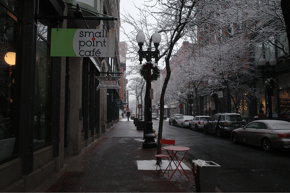 A winter street with parked cars, snow-covered trees, a café sign, and a red table on the sidewalk