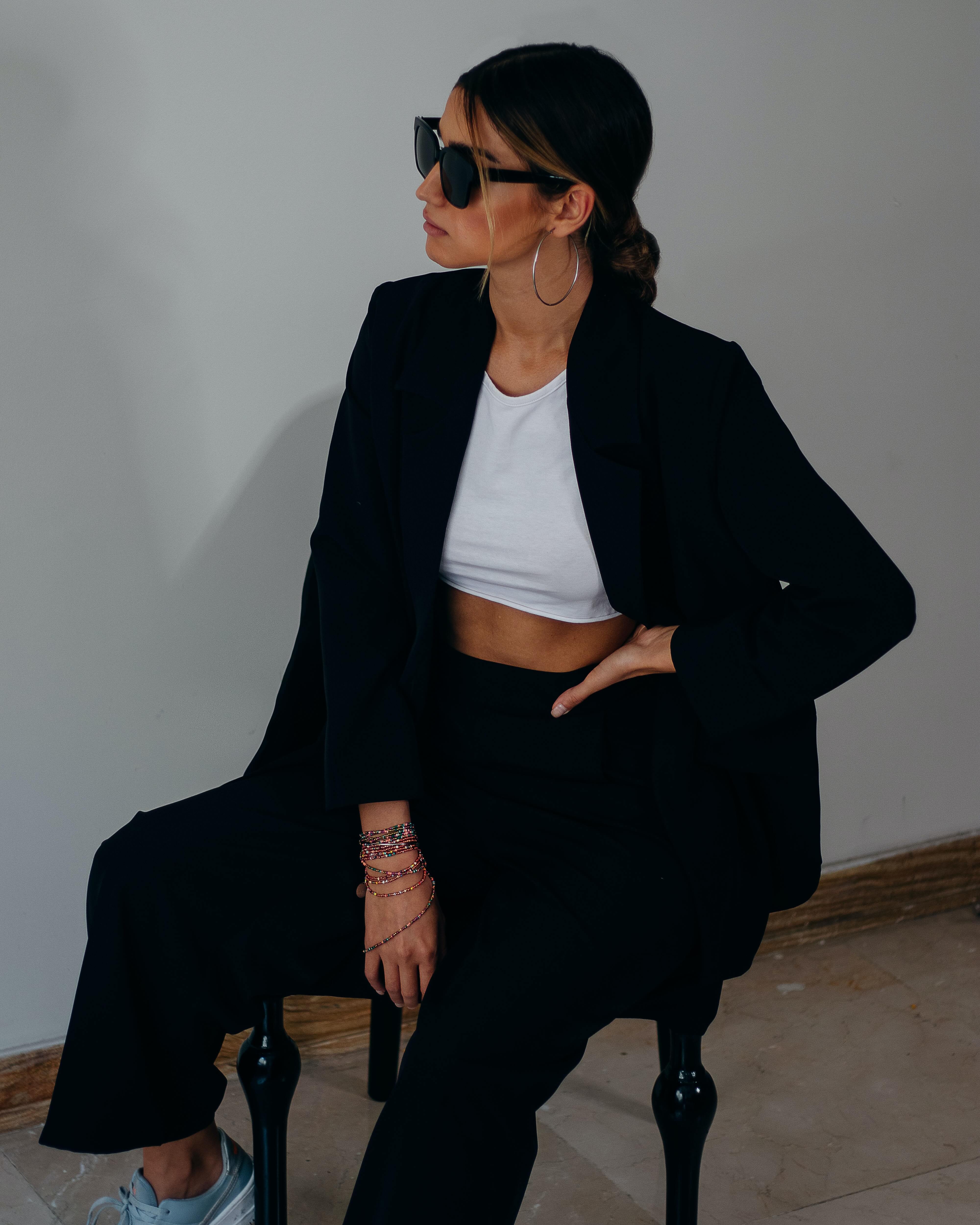Woman wearing black sunglasses, white crop top, and black blazer sitting on a chair with one hand on her hip and colorful bracelets on her wrist.
