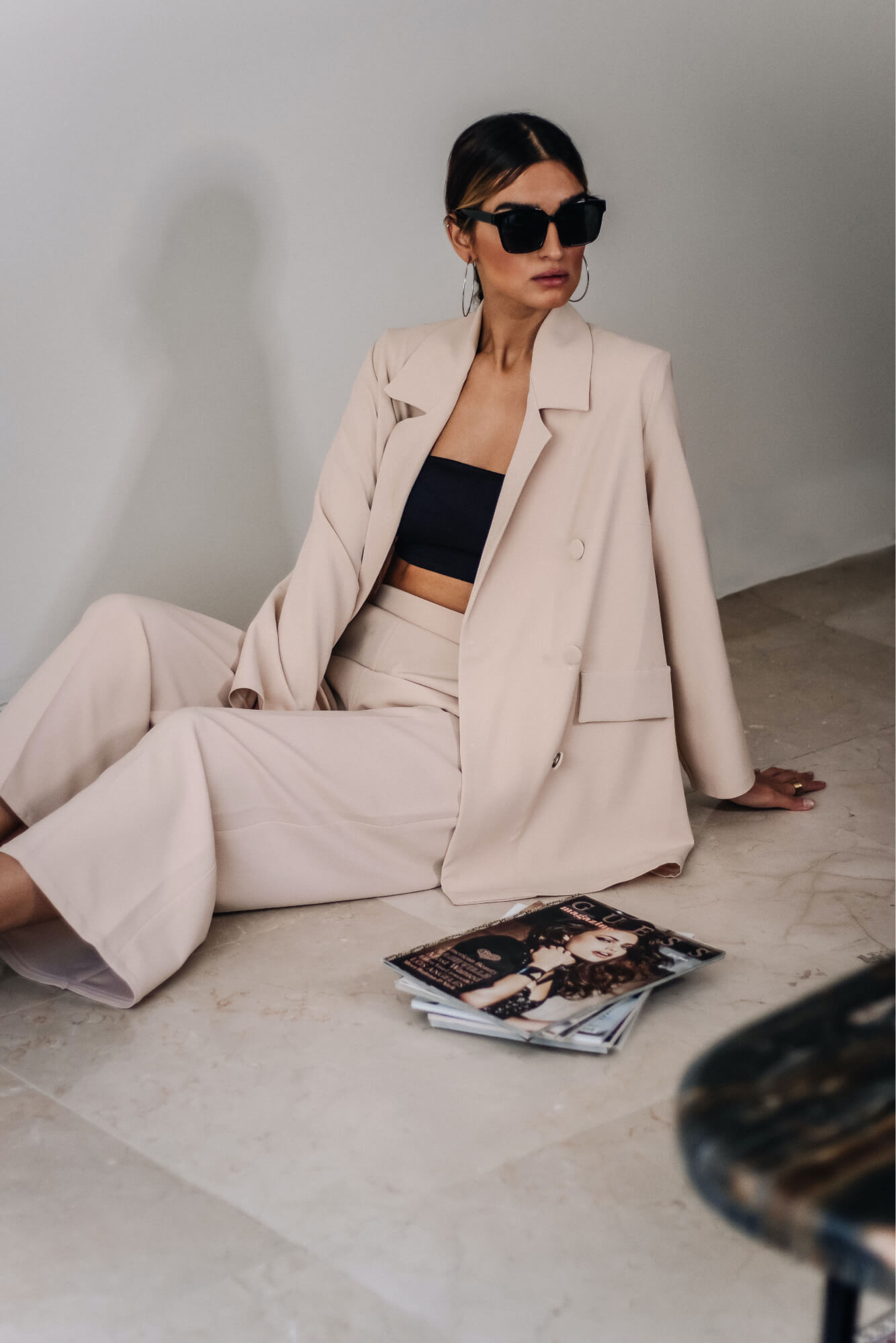 Woman wearing beige suit and black top sitting on the floor next to a stack of magazines.