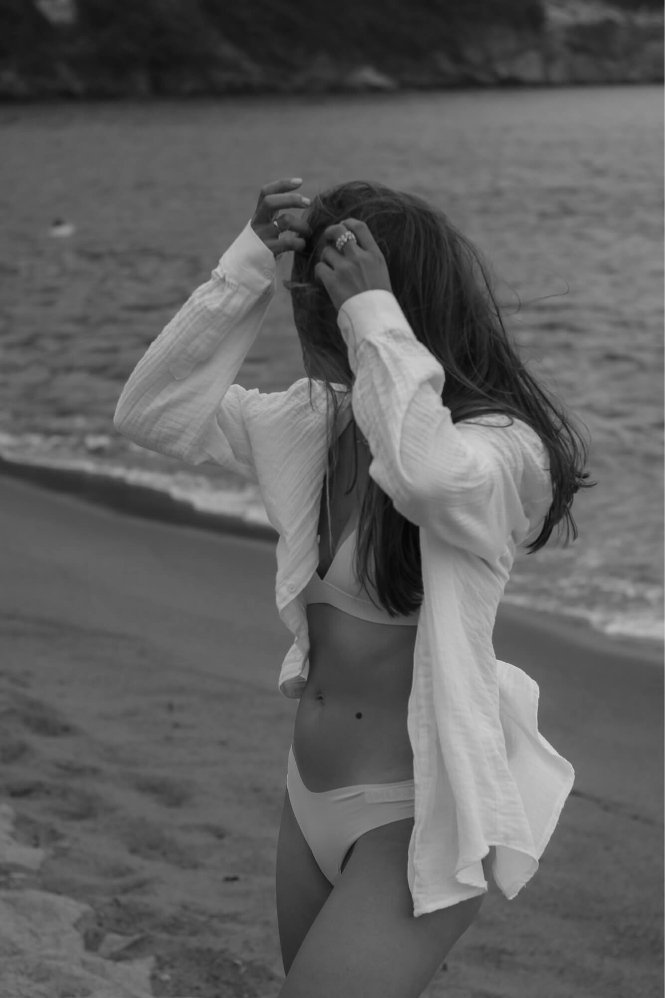 Woman adjusting her hair on a beach, wearing a swimsuit and a loose shirt with the ocean in the background.
