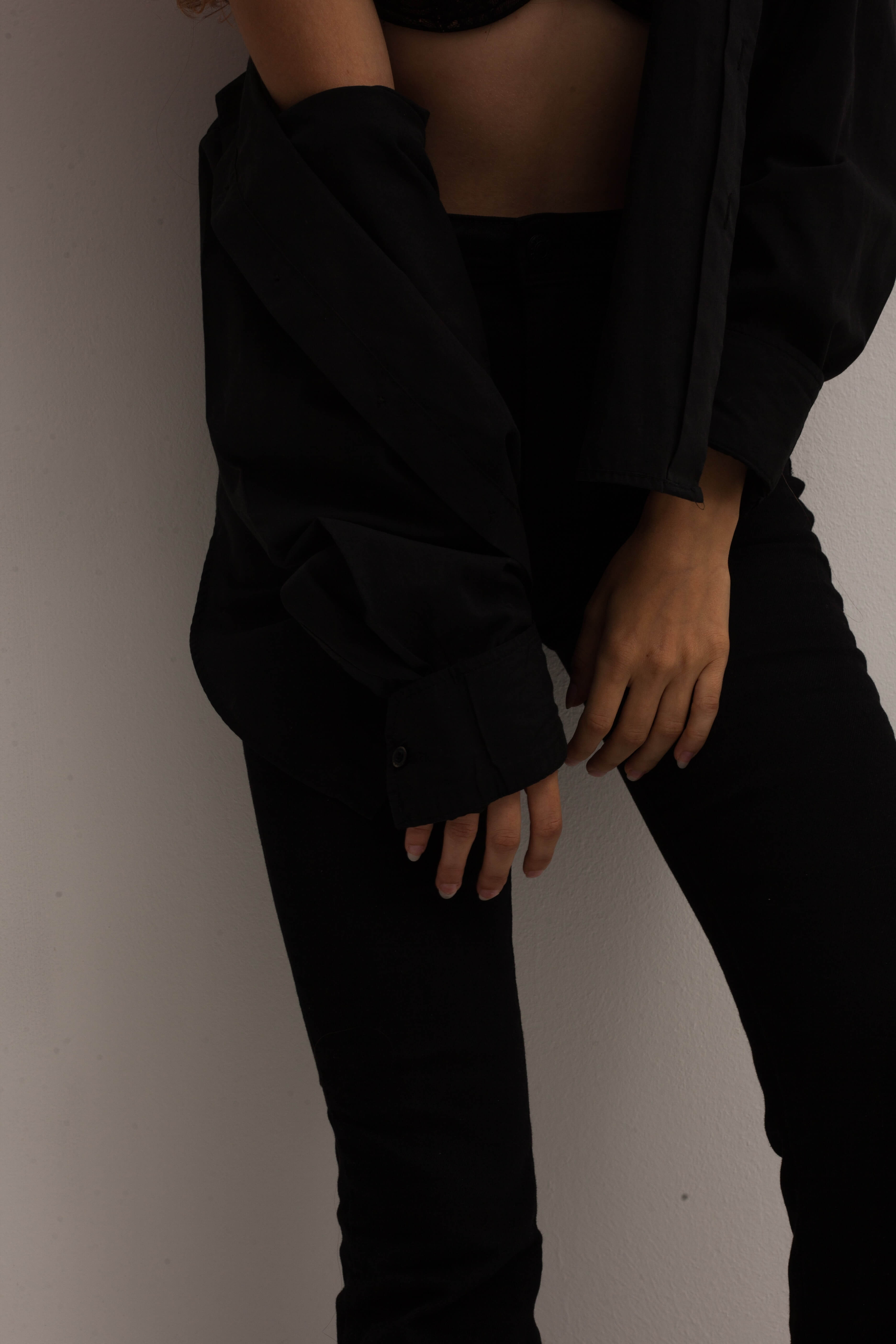 Woman in black high-waisted pants and black shirt with one sleeve draped off shoulder, standing against a plain wall.