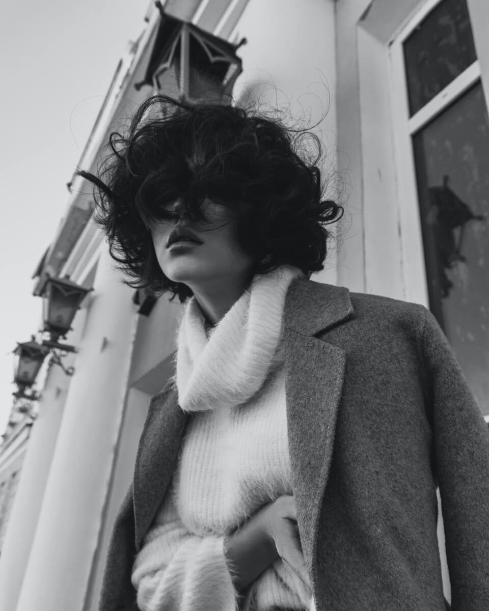 Black and white photo of a person with curly hair obscuring their face, wearing a turtleneck sweater and coat, standing by a building with lanterns.