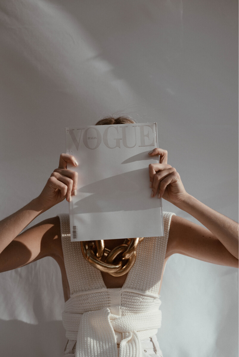 Person in white knitwear and large gold chain holding a Vogue Italia magazine in front of their face.