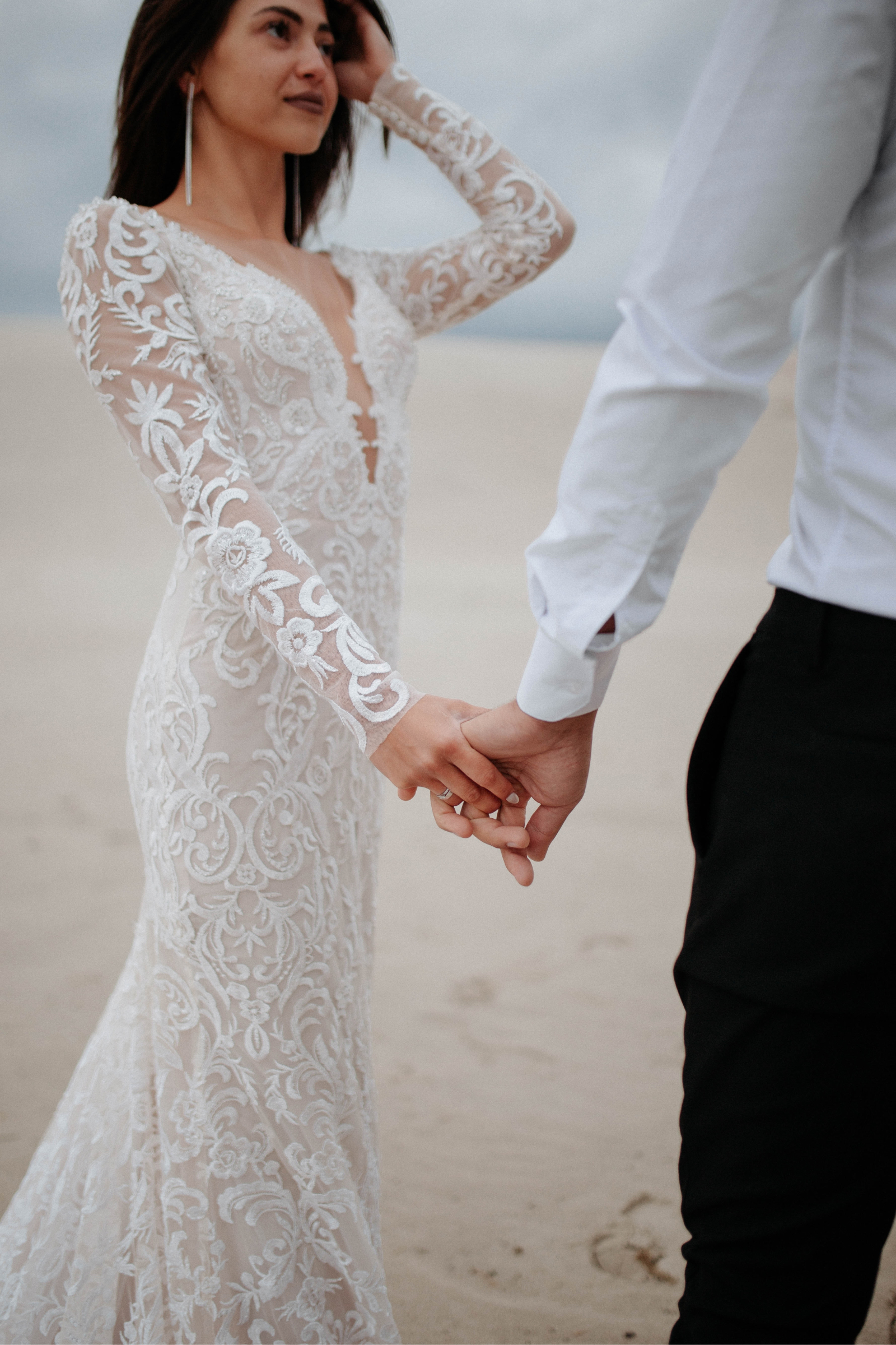 Bride in a white lace wedding dress holding hands with groom wearing a white shirt and black pants on a sandy beach.