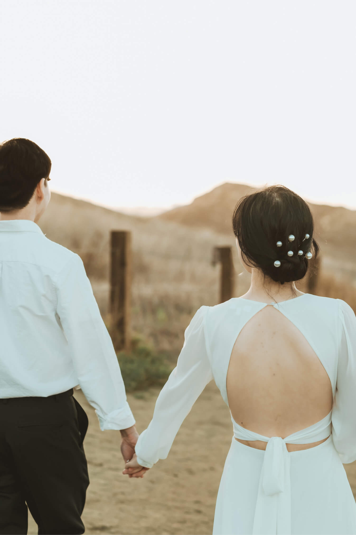 Couple holding hands walking outdoors with the woman in a white dress featuring an open back and pearl hairpins.