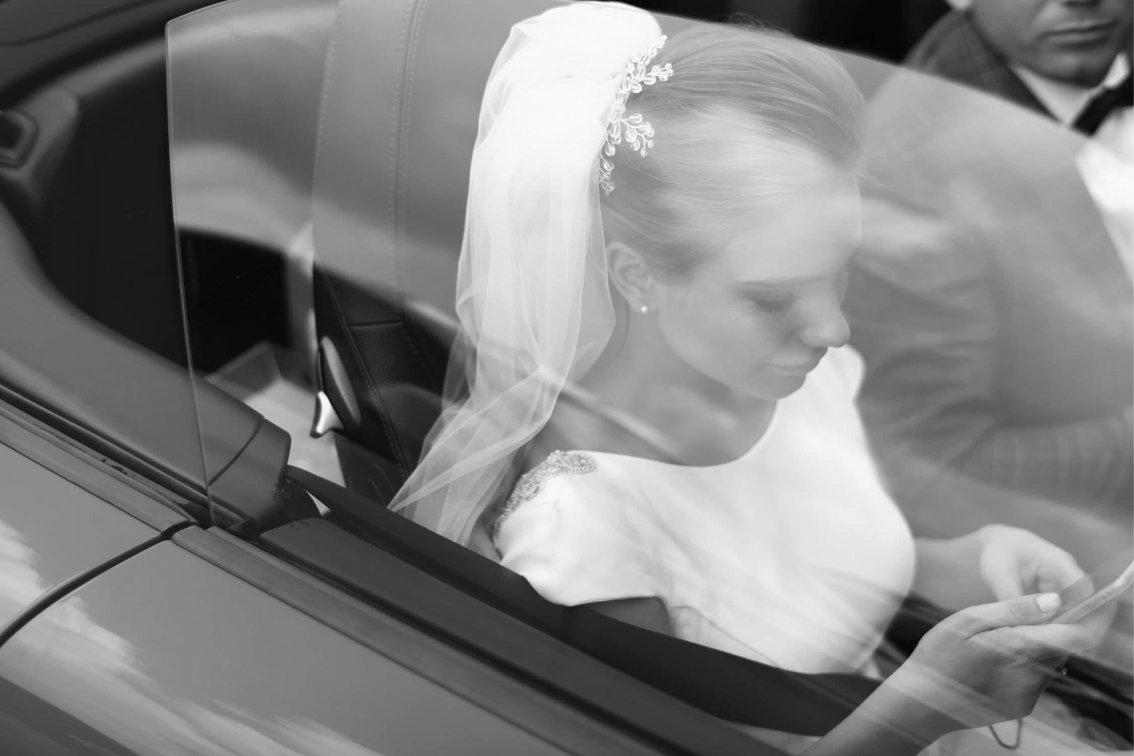 Bride in a wedding dress and veil sitting in a car, looking at a phone.
