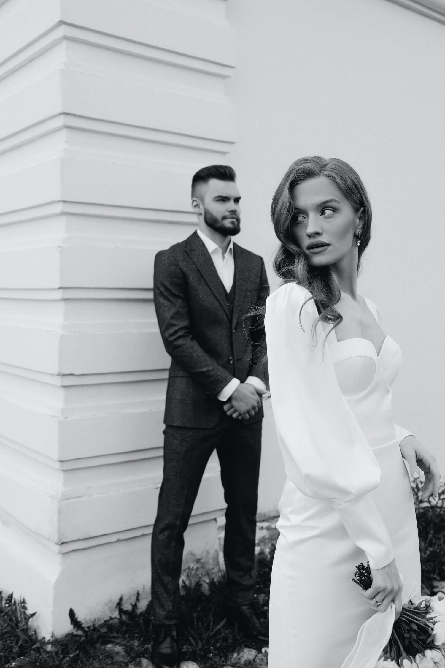 Black and white photo of a bride in a white dress holding flowers looking to the side with a groom in a dark suit standing behind her near a building corner.