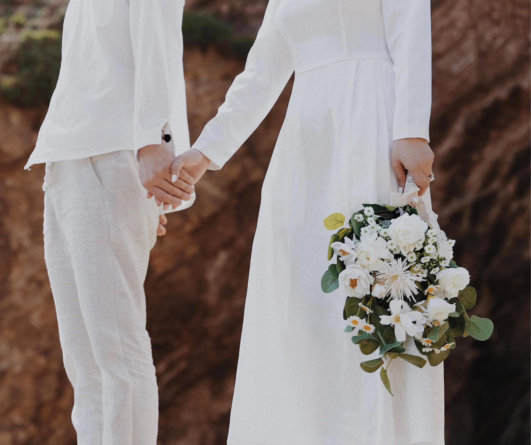 Couple dressed in white holding hands, with one holding a bouquet of white flowers and greenery.