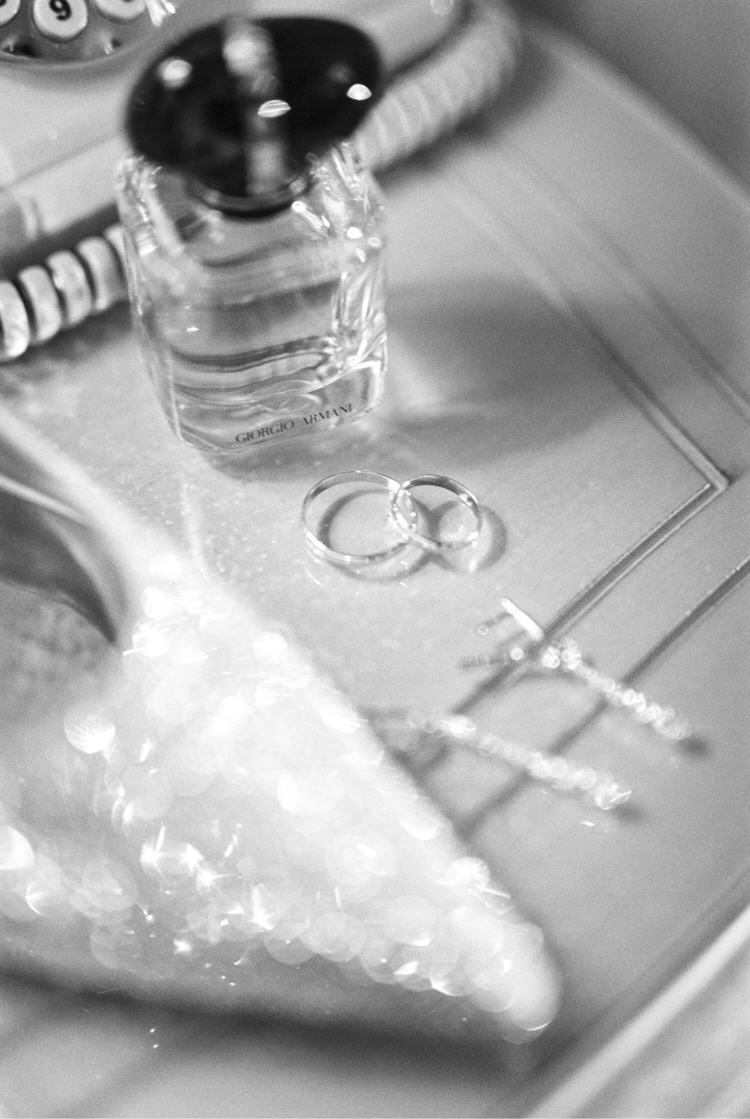 Black and white close-up of a Giorgio Armani perfume bottle, two rings, and blurred sparkling jewelry on a tray.