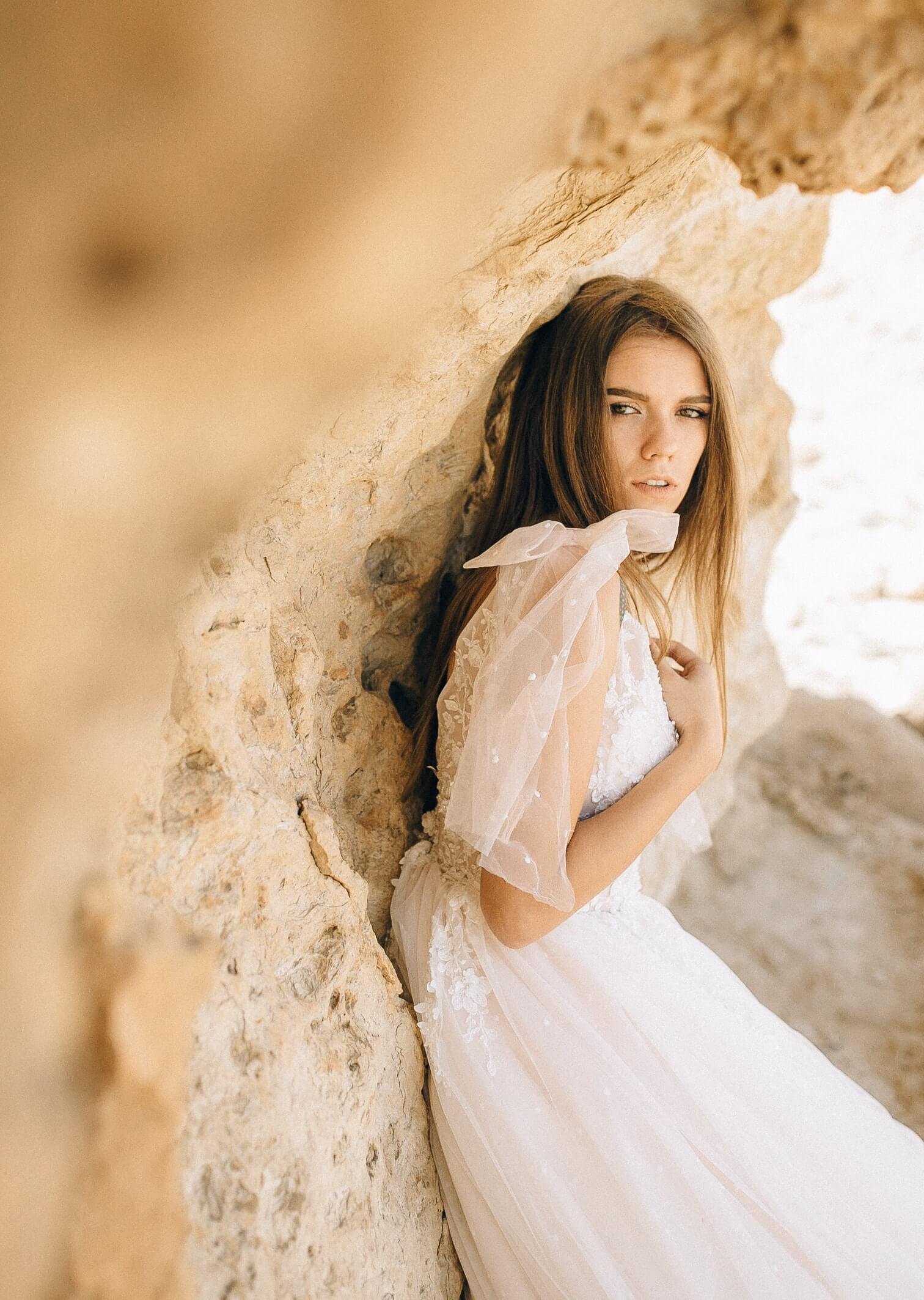 Young woman in a white lace dress leaning against a beige stone wall with a sheer bow on her shoulder.