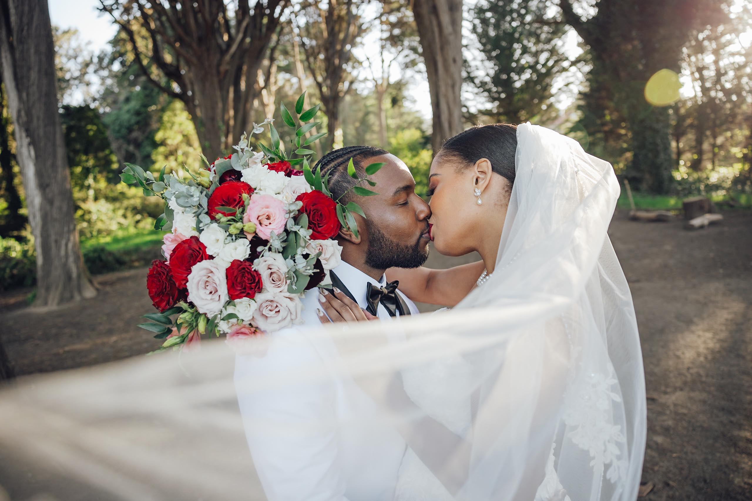Bride and groom sharing a kiss outdoors with the bride's veil flowing and holding a bouquet of red, pink, and white flowers.